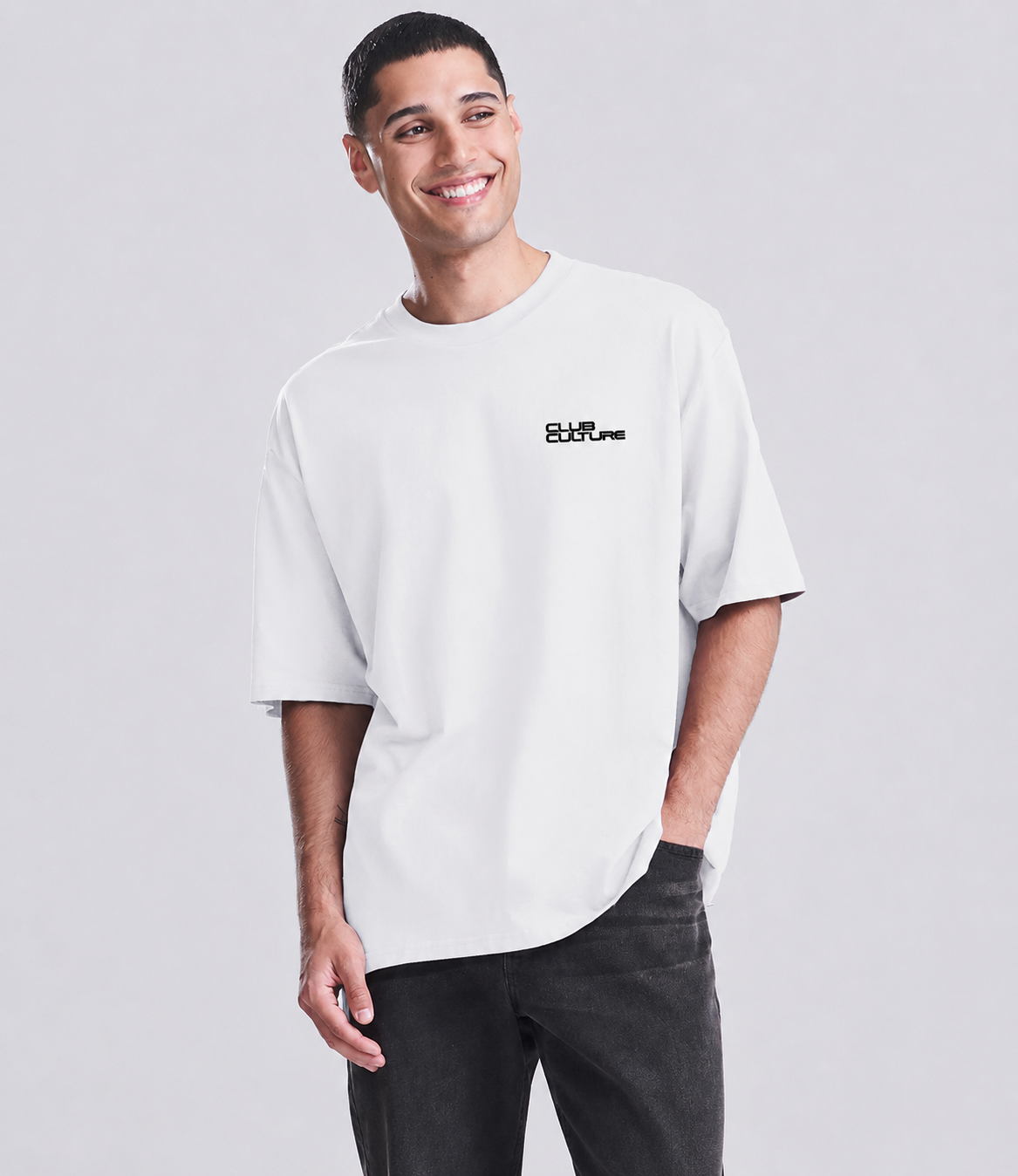 A man wearing a white oversized short-sleeve t-shirt with a small black 'SHIRTS' logo on the left chest, paired with dark gray jeans, against a light gray studio background.
