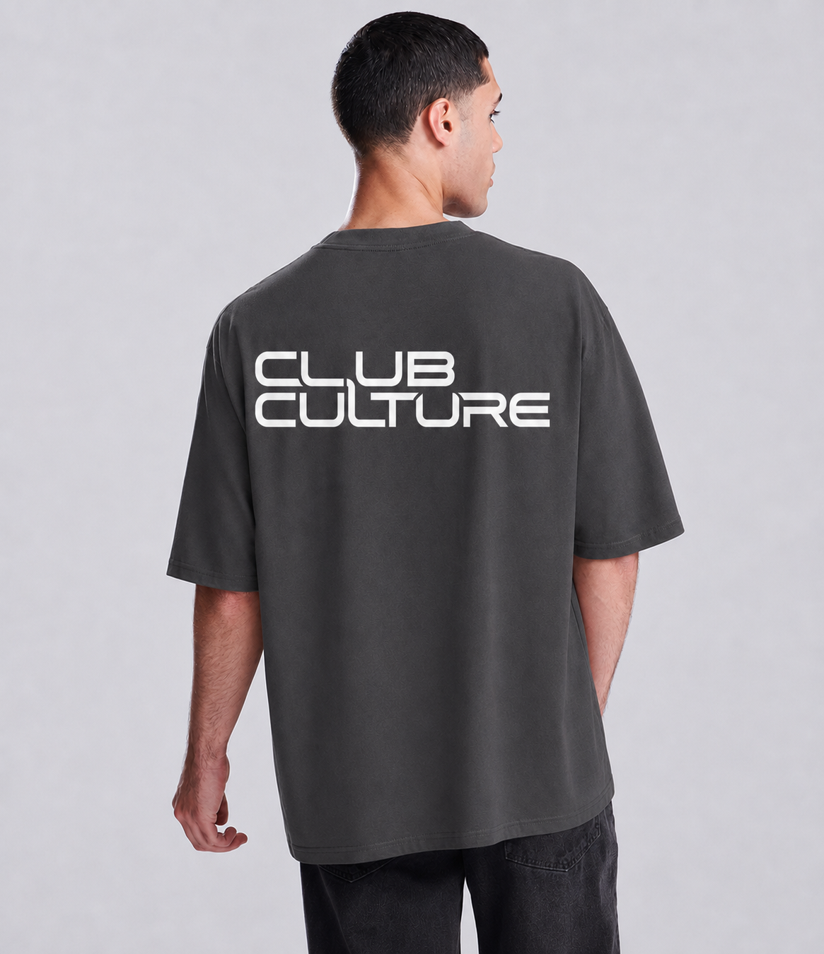 A man viewed from the back wearing a dark charcoal gray oversized t-shirt with 'CLUB CULTURE' printed in white across the upper back, paired with dark pants against a light gray studio background.
