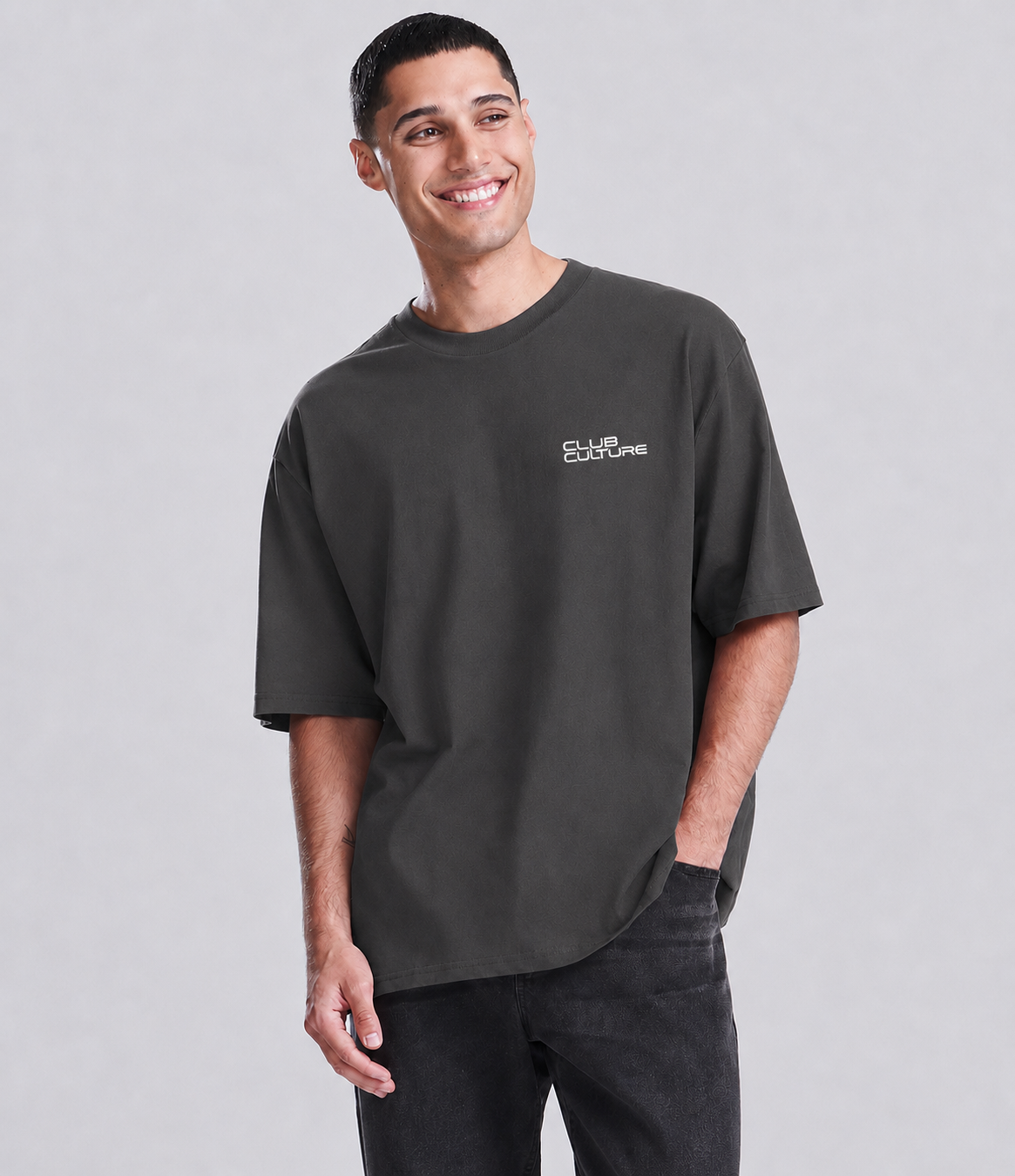 A man wearing a dark charcoal gray oversized t-shirt with a small white 'School of Crime' logo on the left chest, paired with black jeans, against a solid light gray background.
